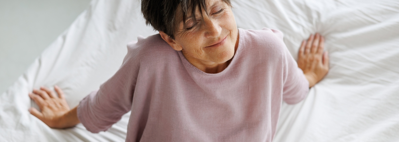 Mature woman lying back on white bedsheets with eyes closed, happy